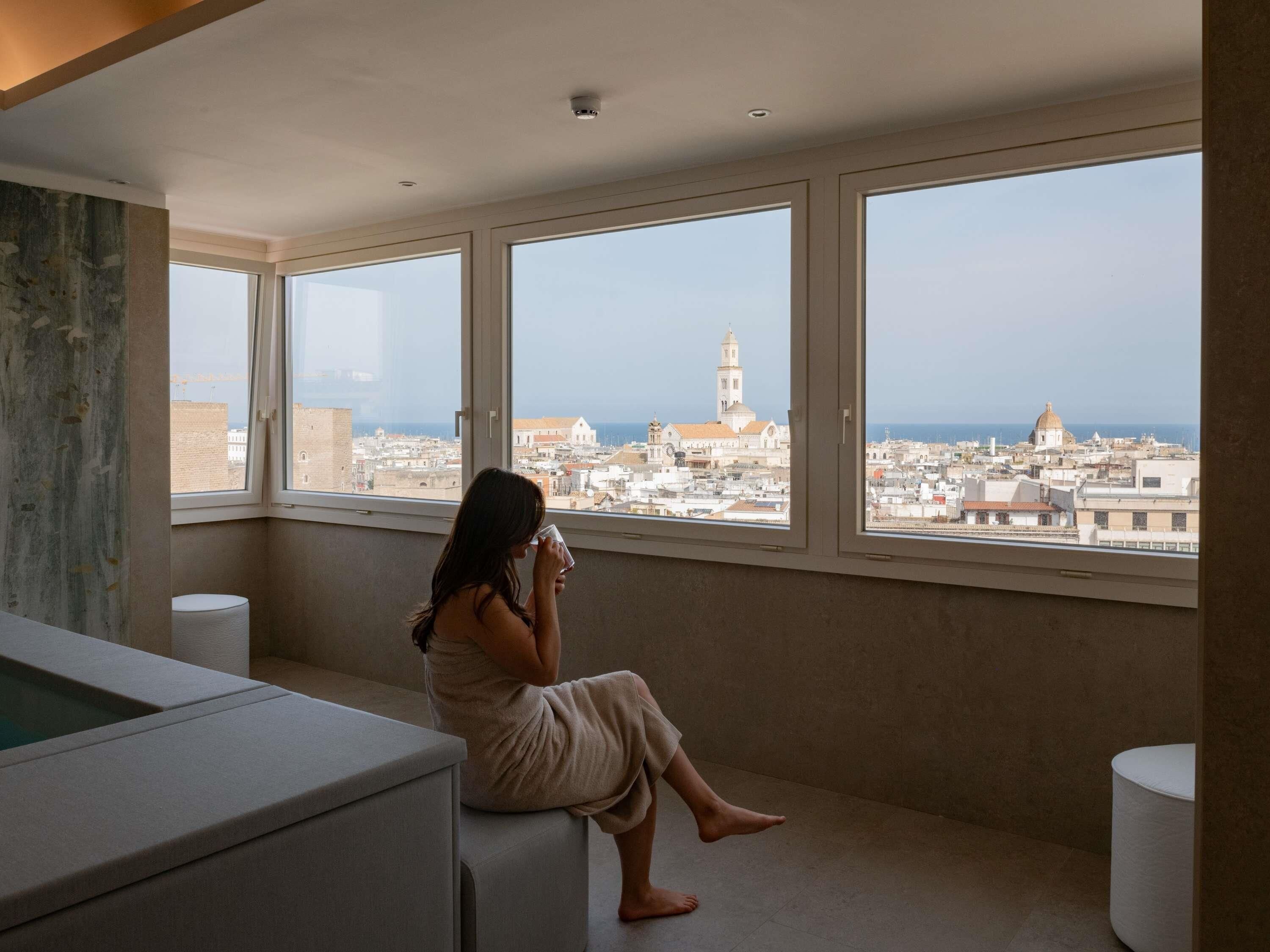 A room with a large window overlooking a cityscape, a bathtub, and a toilet.