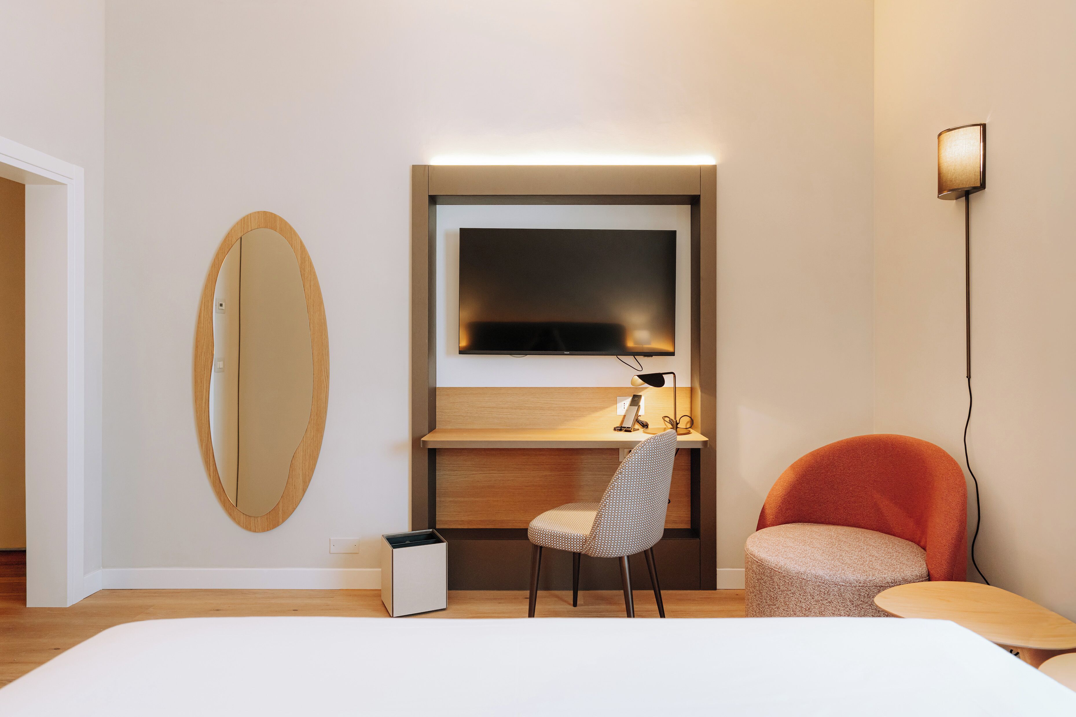 A modern hotel room with a wooden desk, a flat-screen TV, a round mirror, a red chair, and a small table.