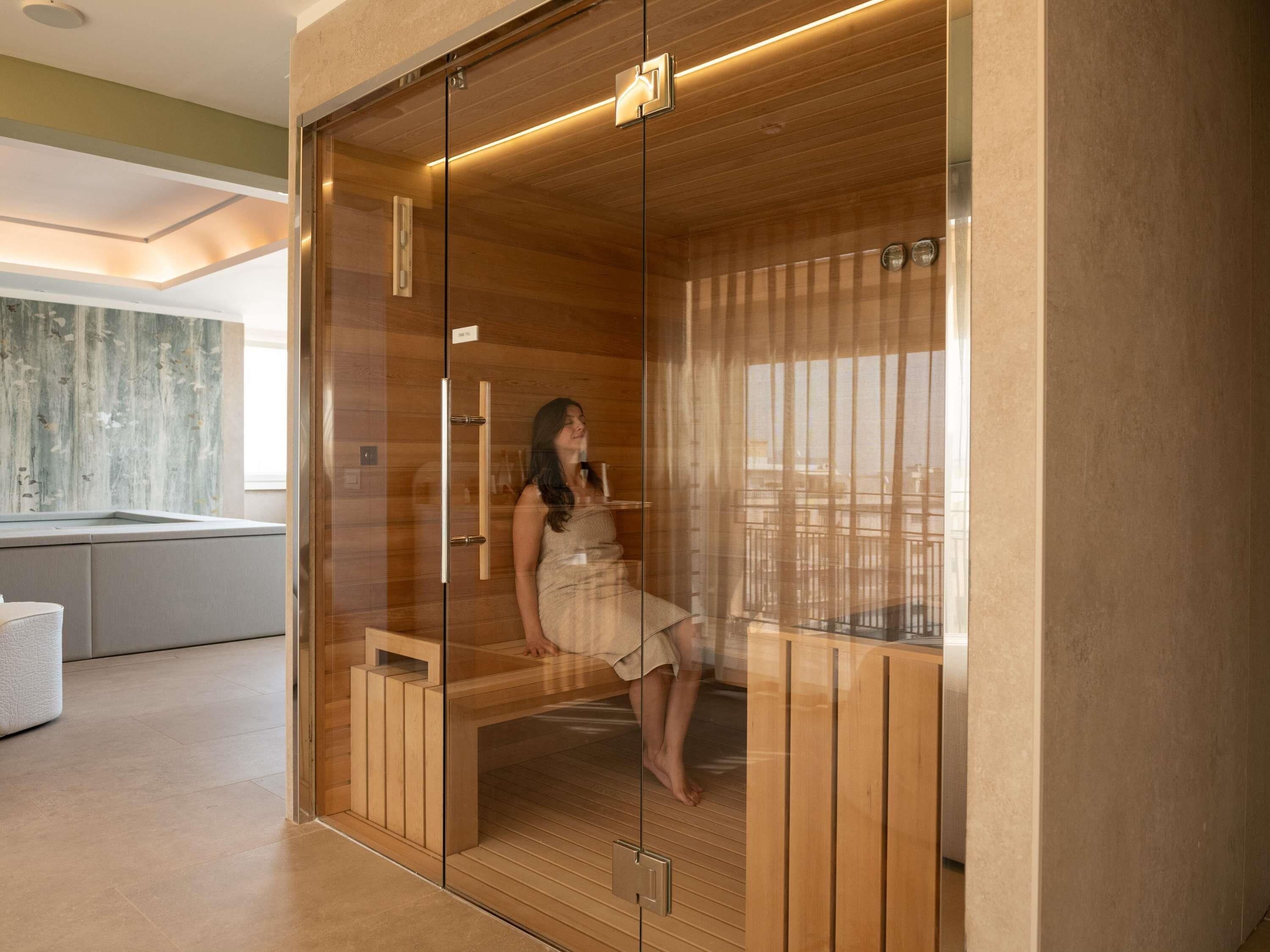 A modern sauna with a glass enclosure, wooden interior, and a view of a bathroom with a bathtub.