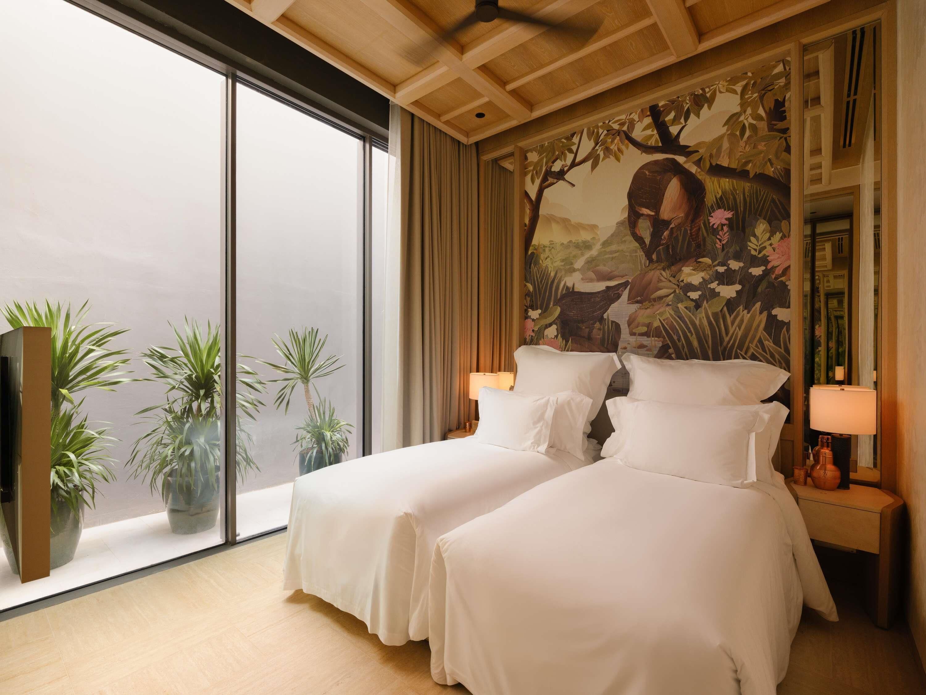 Une chambre d’hôtel avec deux lits, une grande fenêtre, un ventilateur de plafond et une fresque murale.