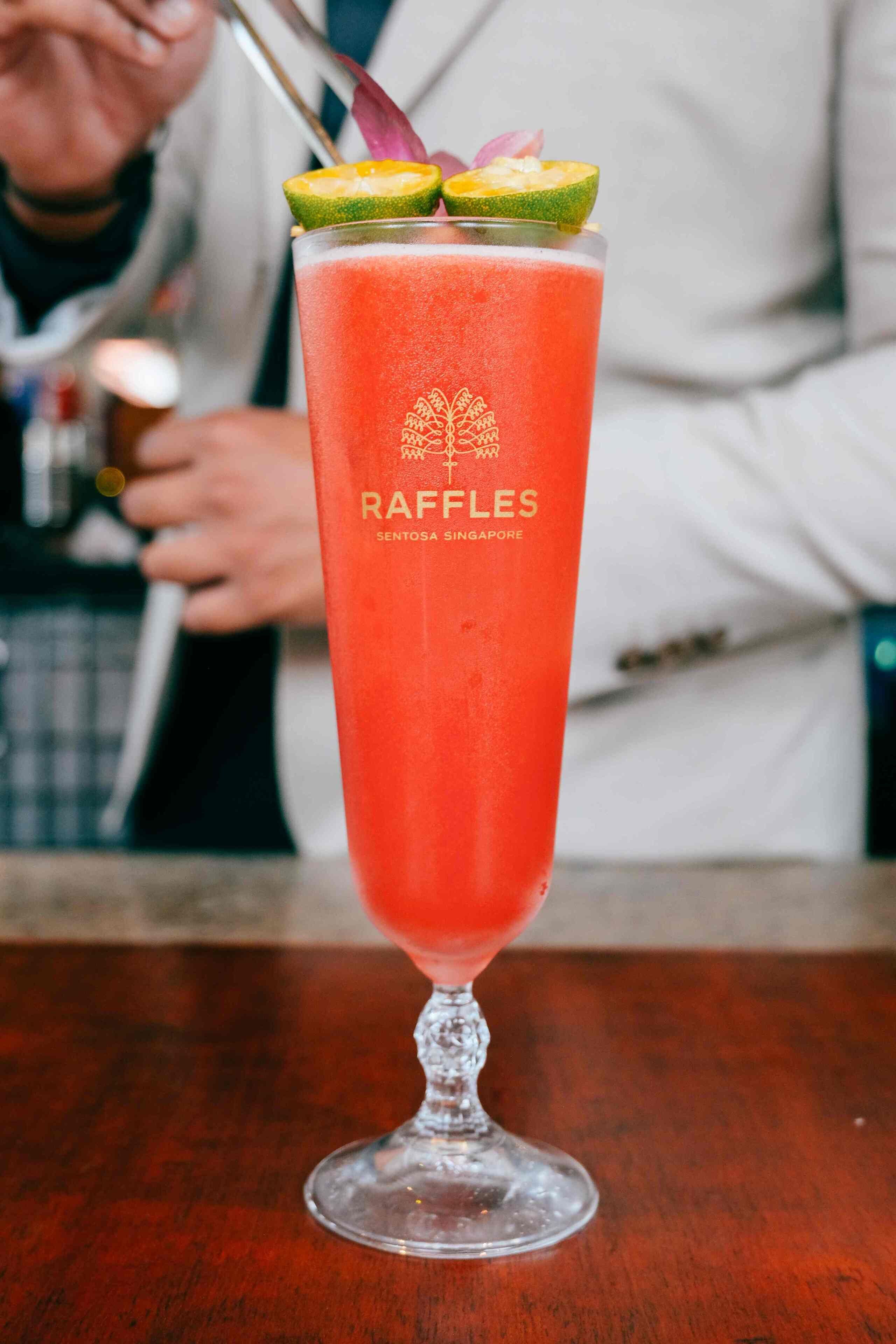 A tall glass of red beverage with a lime garnish, labeled "RAFFLES SENTOSA 5 SINGAPORE."