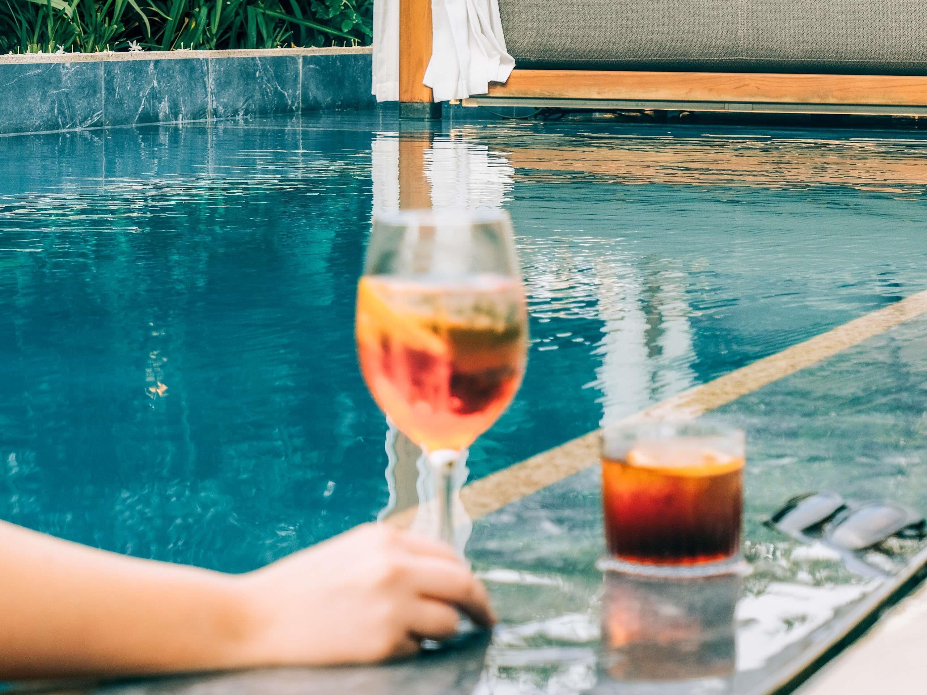 A glass of wine and a cocktail on a table by a pool.