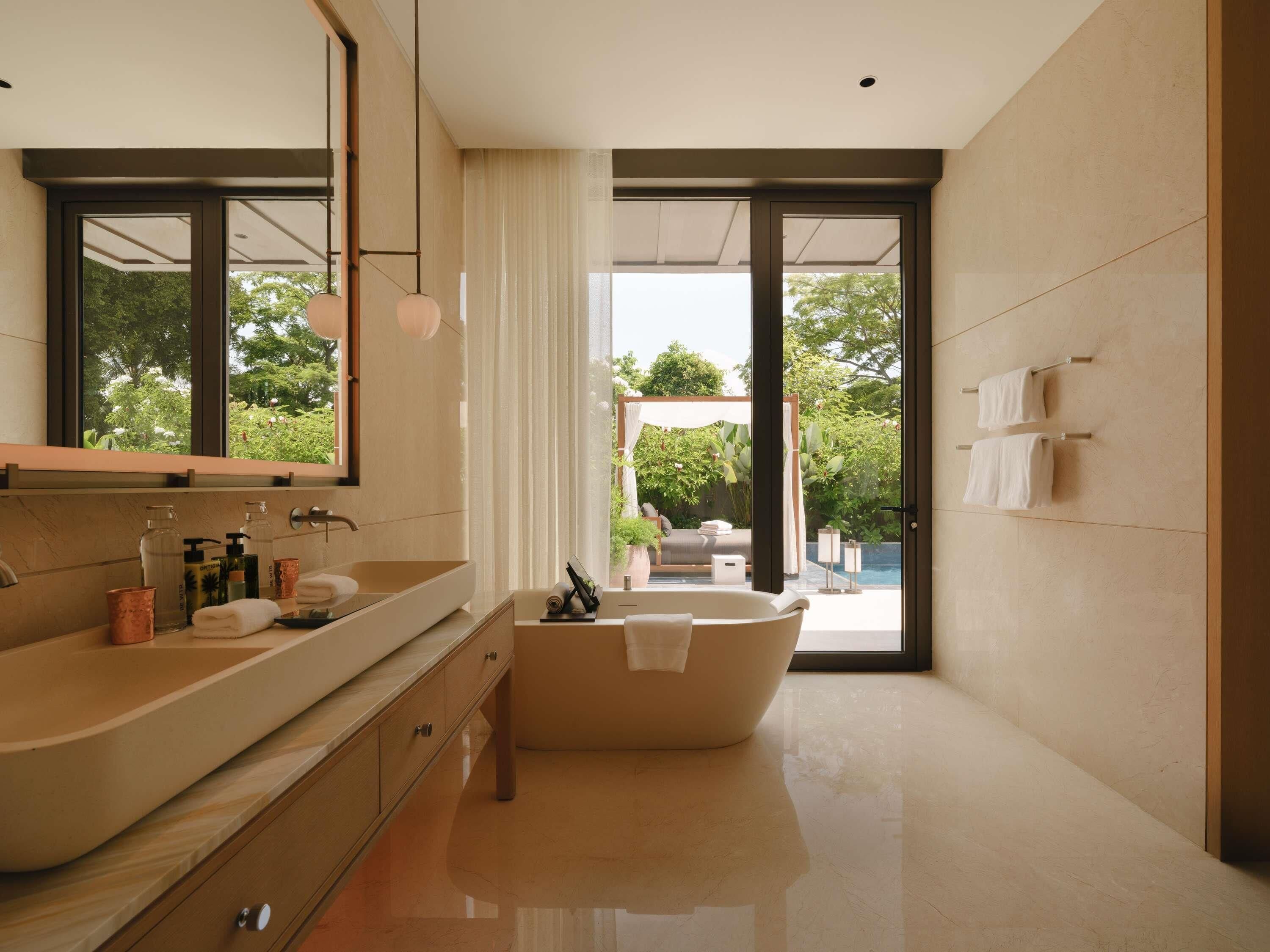 A modern bathroom with a large bathtub, a double vanity with a mirror, and a view of a pool and garden.