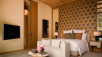 A bedroom with a bed, a sofa, a round coffee table with books and flowers, a TV, and a decorative wall panel.