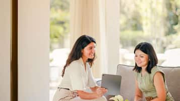 Two people sitting on a couch, one holding a tablet, in a room with a large window and a plant on the table.