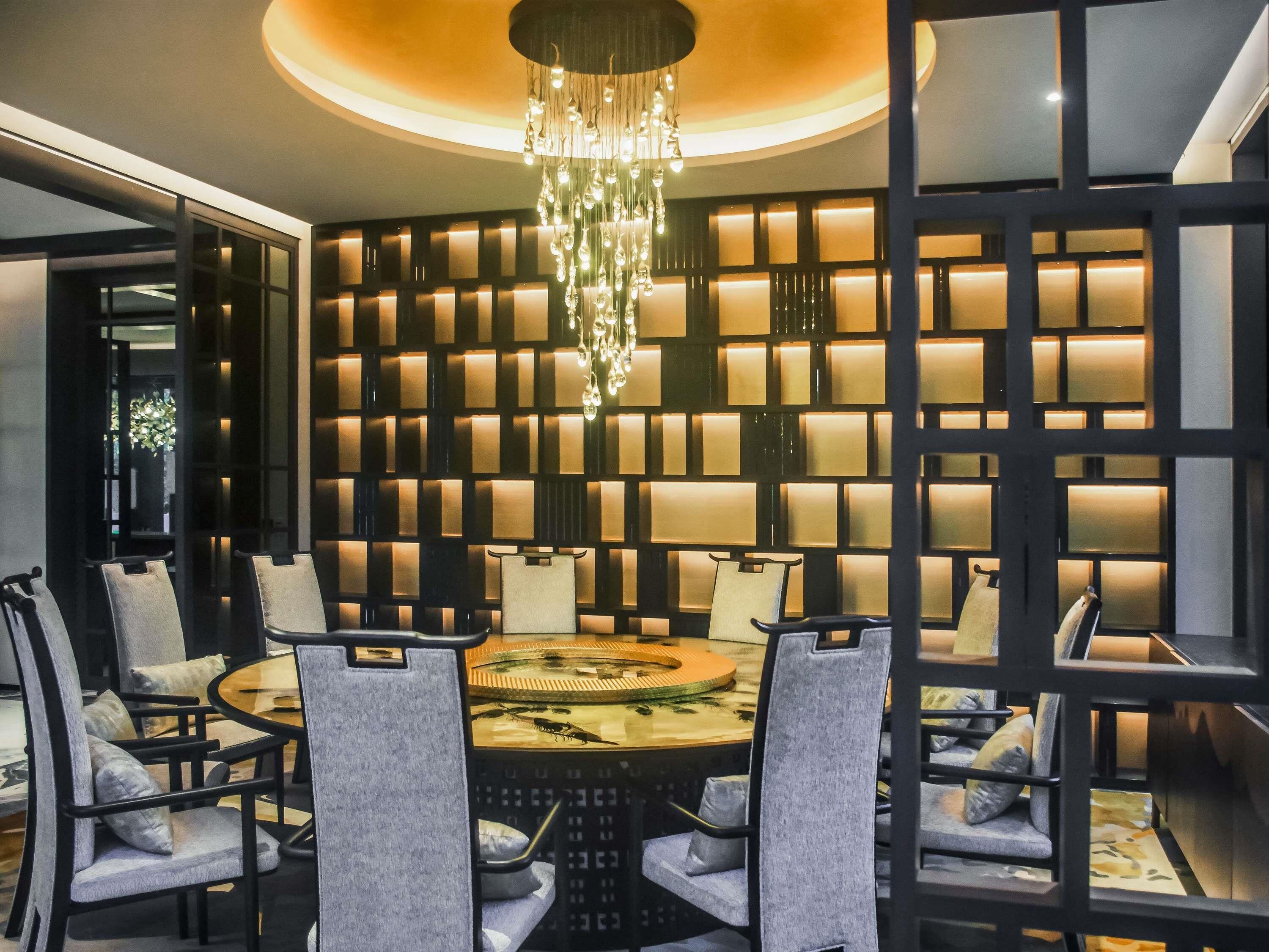 A modern dining area with a round table, multiple chairs, a chandelier, and a wall with illuminated panels.