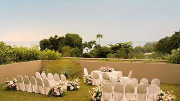 An outdoor wedding setup with white chairs and tables, surrounded by greenery and a view of the ocean.
