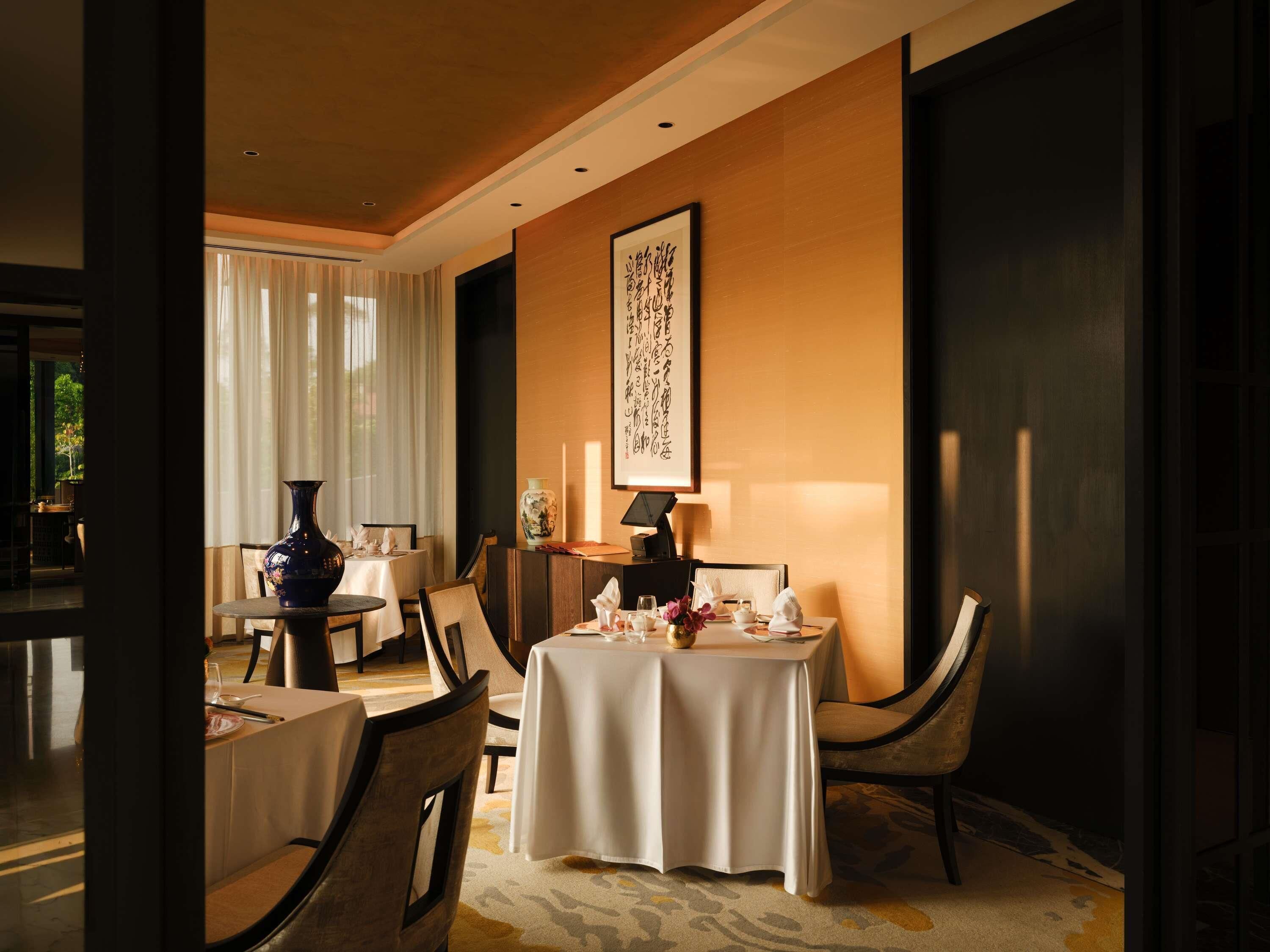 A dining area with tables set for two, a vase, and a framed calligraphy piece on the wall.