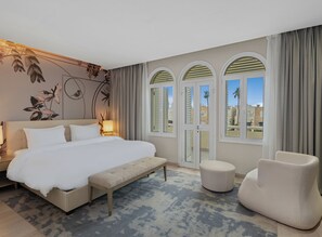 A modern hotel room with a large bed, a bench, a chair, and a view of buildings through arched windows. - Rixos Obhur Jeddah Resort & Villas (Jeddah)