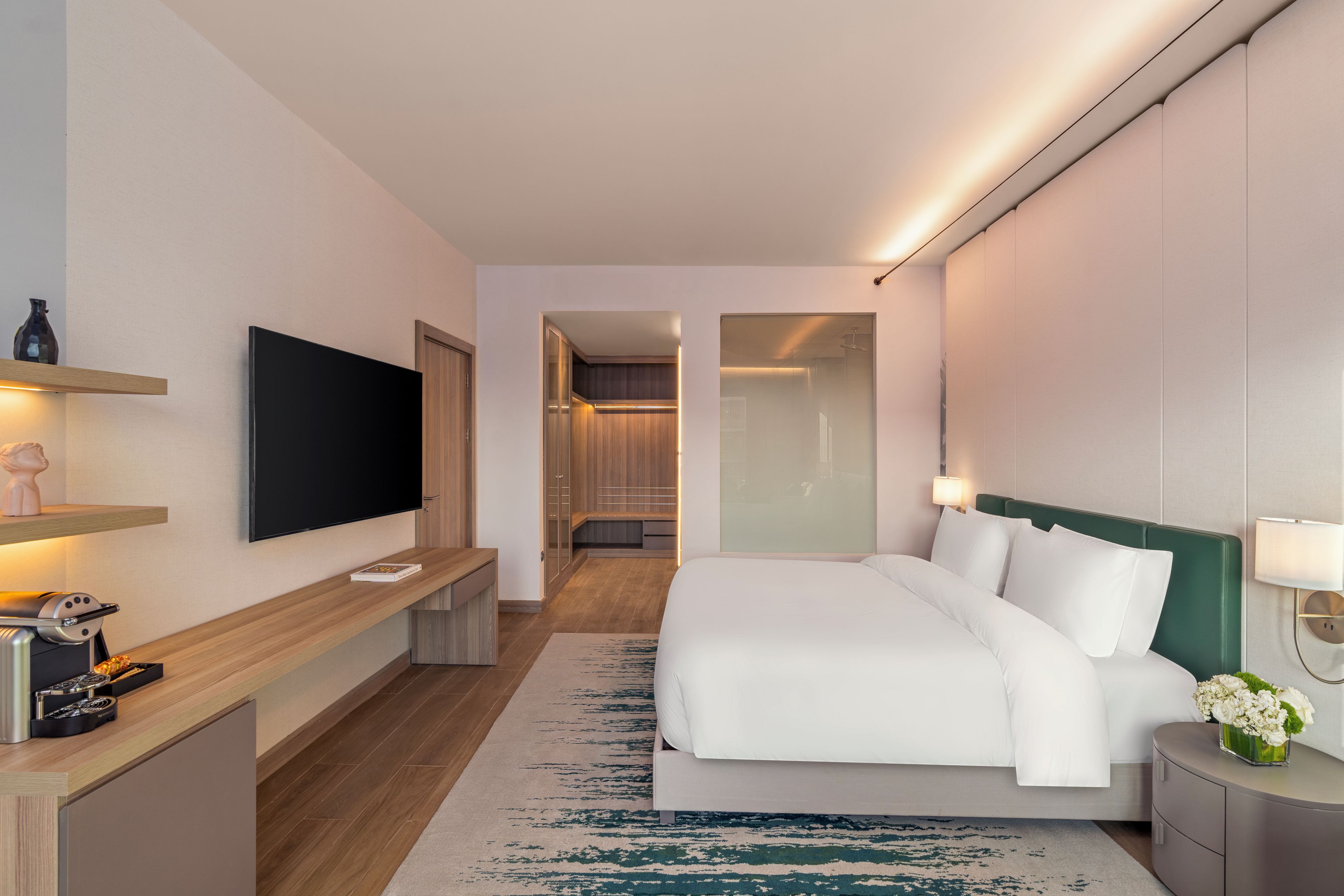 Une chambre d’hôtel moderne équipée d’une télévision à écran plat, d’une table de chevet avec une lampe, d’un lit avec une literie blanche et d’une petite table d’appoint agrémentée d’un vase de fleurs.