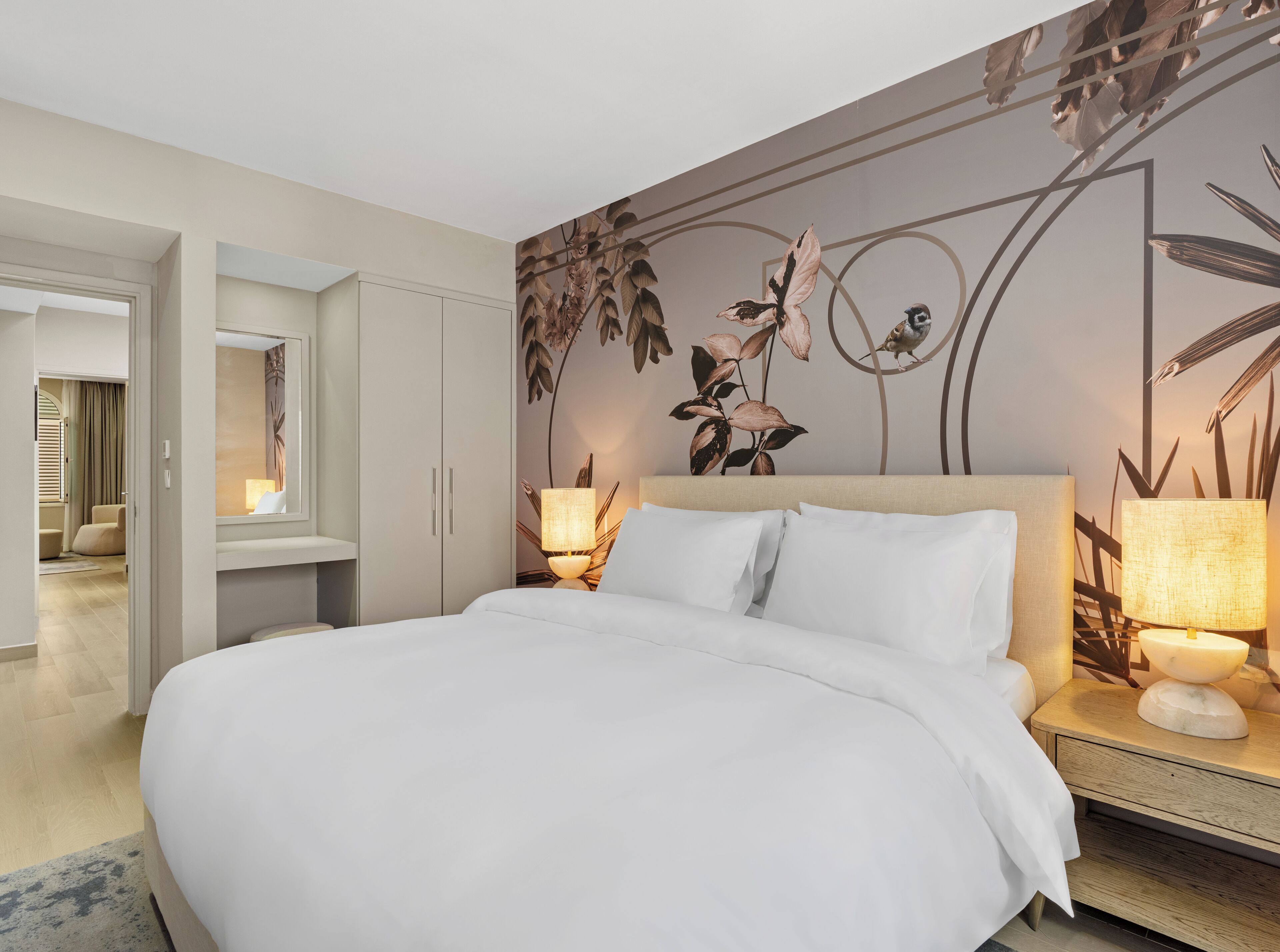 Habitación de hotel con una cama grande, dos mesitas de noche con lámparas, un mural en la pared y una puerta que da a otra habitación.