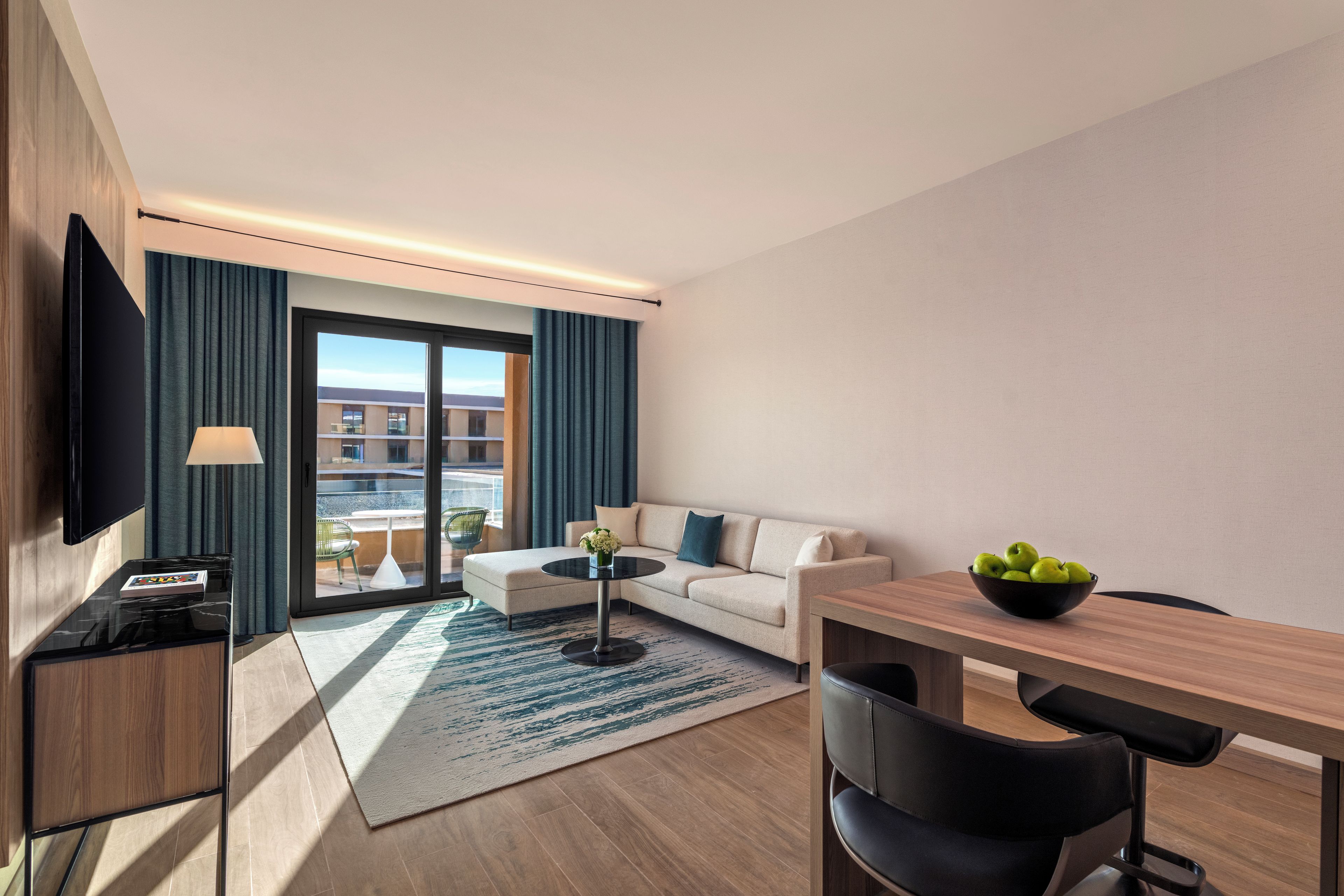 A modern hotel room with a sofa, a dining table, a TV, and a view of the cityscape.