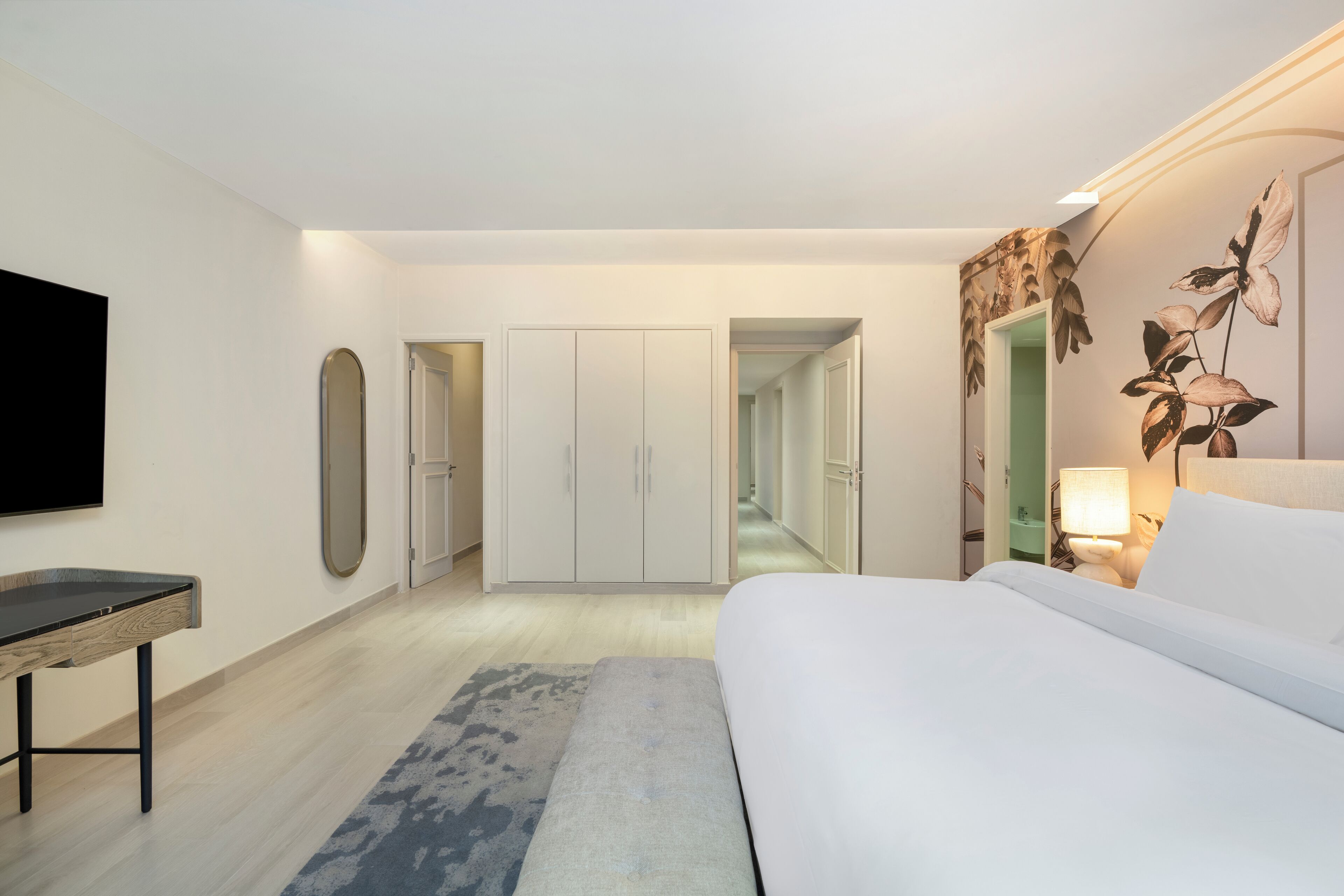 A modern hotel room with a bed, a desk, a flat-screen TV, a wardrobe, and a hallway leading to another room.
