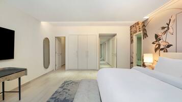 A modern hotel room with a bed, a desk, a flat-screen TV, a wardrobe, and a hallway leading to another room.