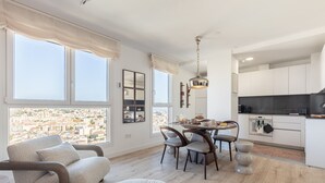 En moderne stue med en sofa, et spisebord med stole og udsigt over byen gennem store vinduer.