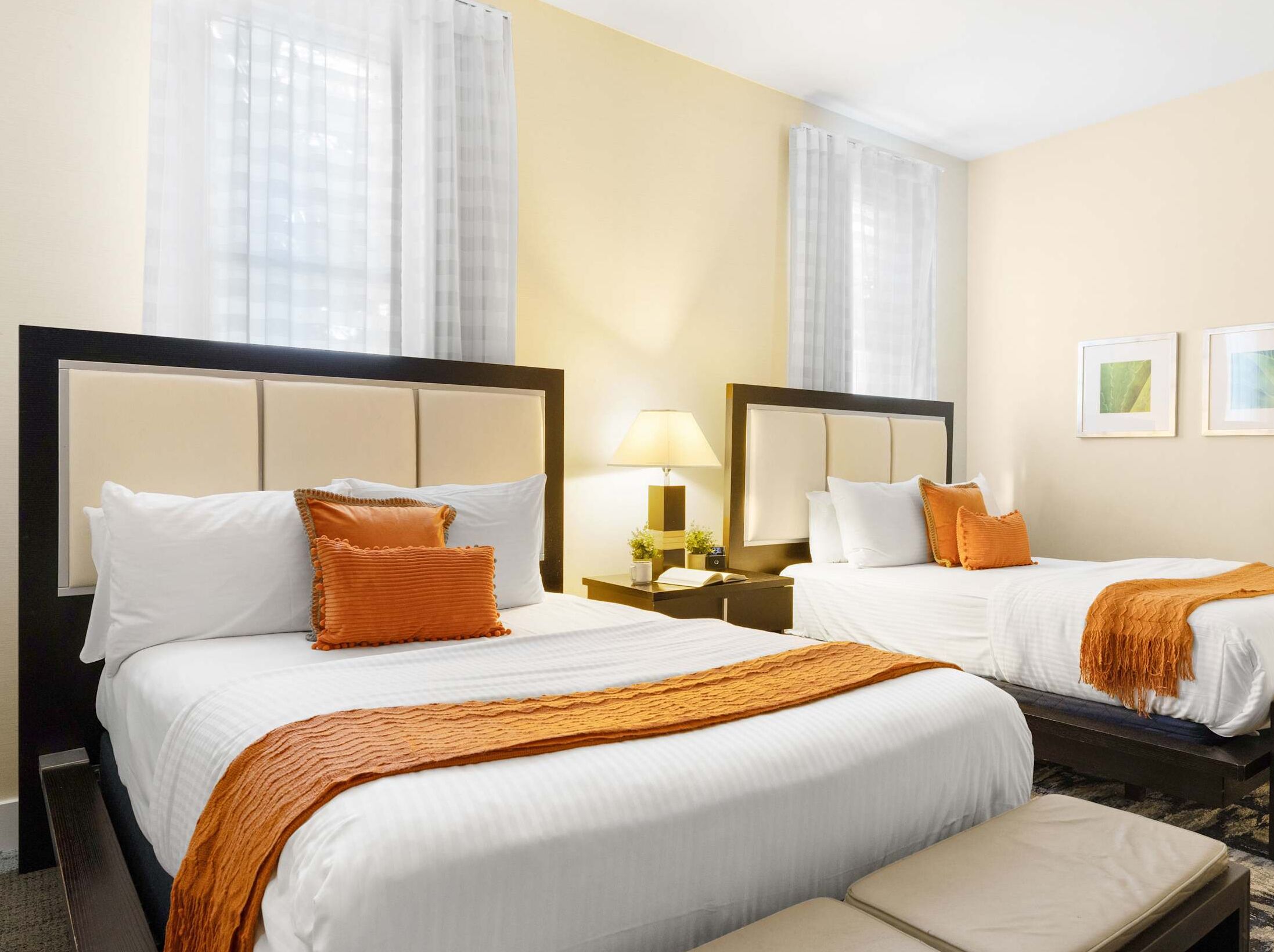 A hotel room with two beds, each with white linens and orange accents, a bedside table with a lamp, and a small plant.