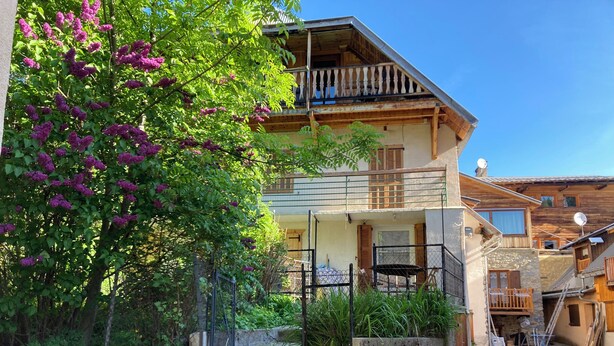 Un bâtiment à deux étages doté d’un balcon, d’un escalier en pierre et d’un jardin où poussent des fleurs violettes.