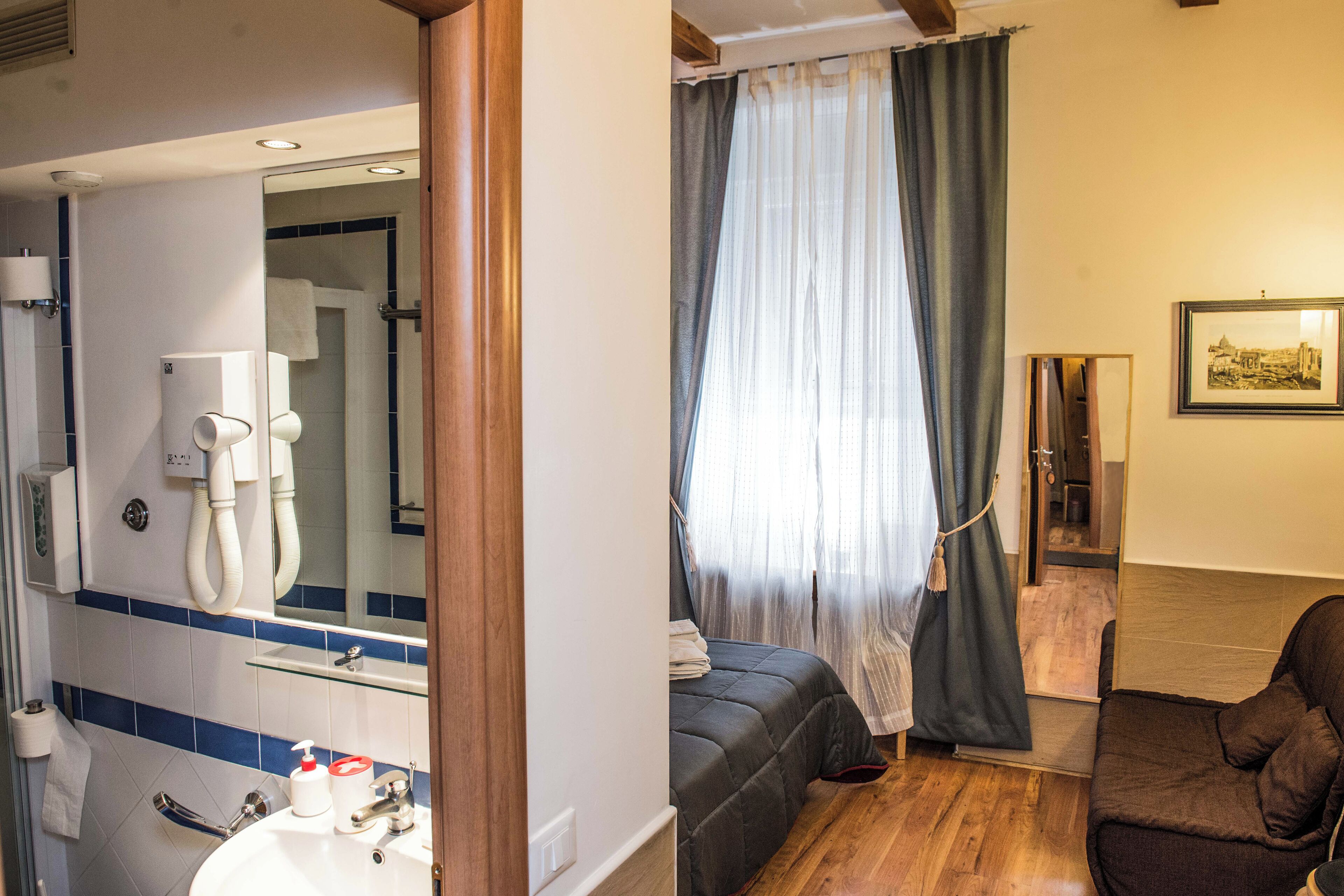 Baño con lavamanos, espejo y toallero. Cuenta con zona de ducha con puerta de cristal y un inodoro. El piso es de madera y la cama tiene una colcha oscura.