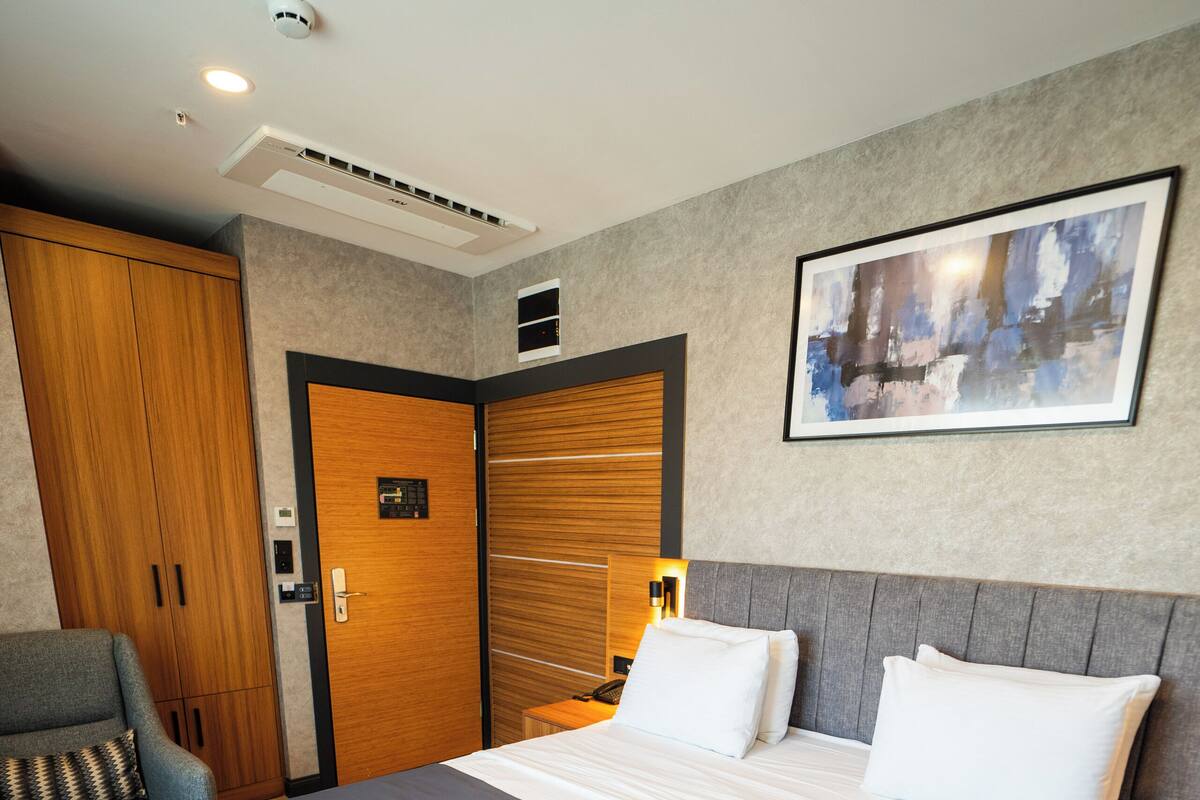 A hotel room with a bed, a chair, a wardrobe, a painting on the wall, and a ceiling with lights and an air conditioner.