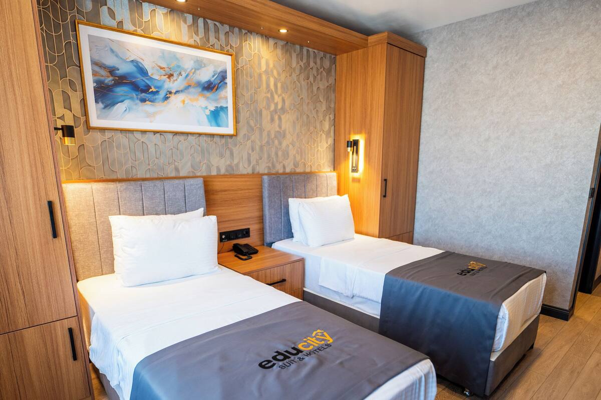 A hotel room with two beds, a wooden headboard, a painting, and a nightstand with a lamp.