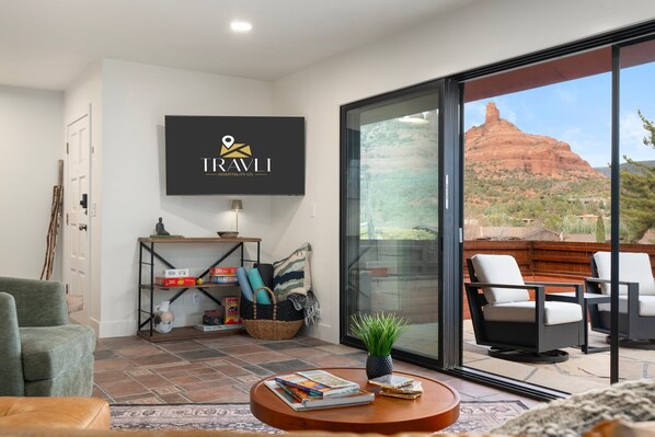 A modern living room with a large TV displaying the TRAVLI logo, a comfortable seating area with a view of a red rock formation through large glass doors.