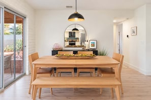 A dining area with a wooden table, chairs, a central island with a bowl of fruit, a black pendant light, and a large mirror above the island.