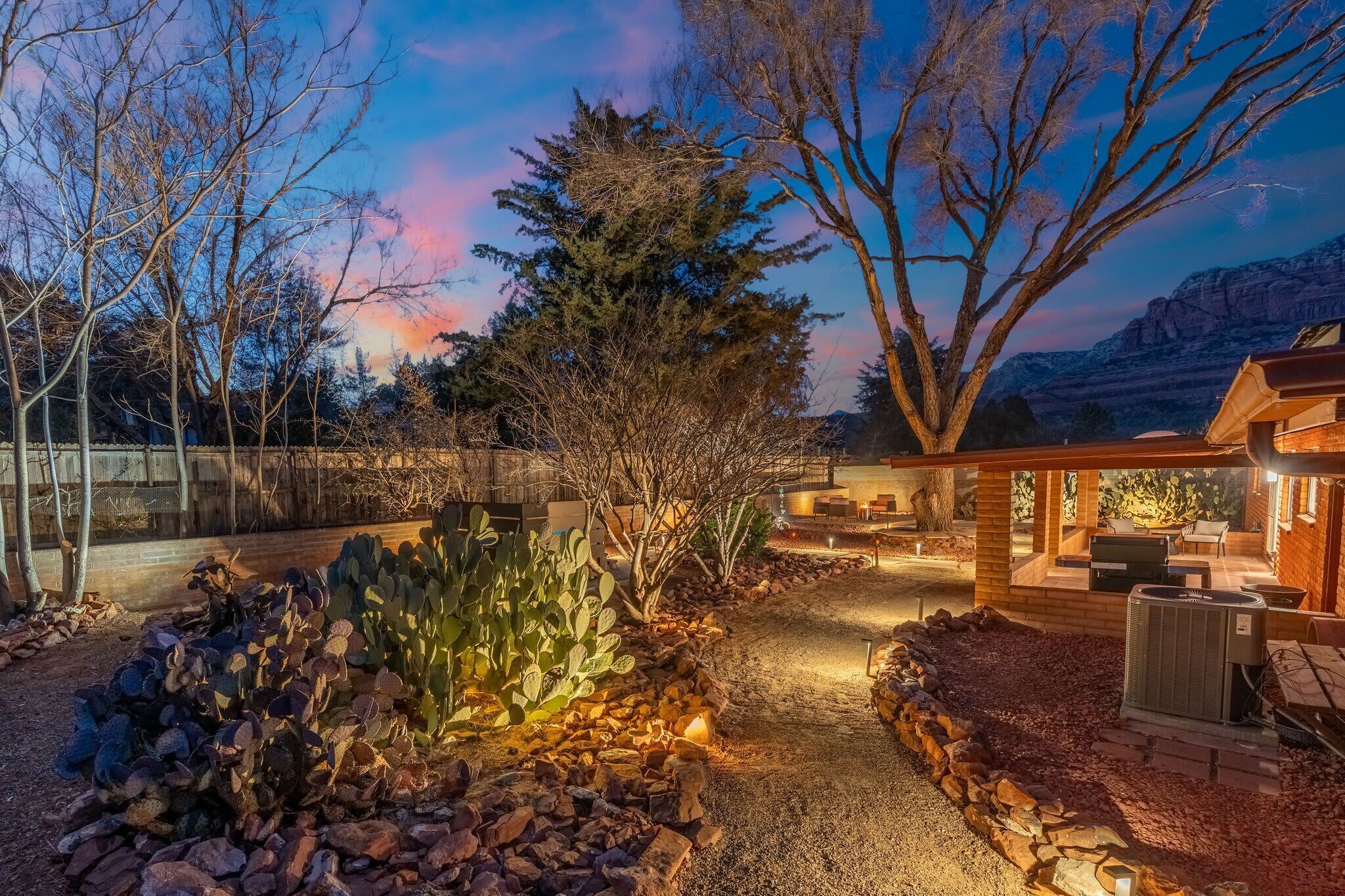 A well-lit outdoor patio area with a stone pathway, surrounded by cacti and trees, leading to a wooden structure with outdoor seating.