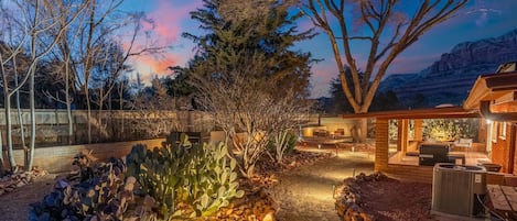 Un patio exterior bien iluminado con un sendero de piedra, rodeado de cactus y árboles, que conduce a una estructura de madera con asientos al aire libre.