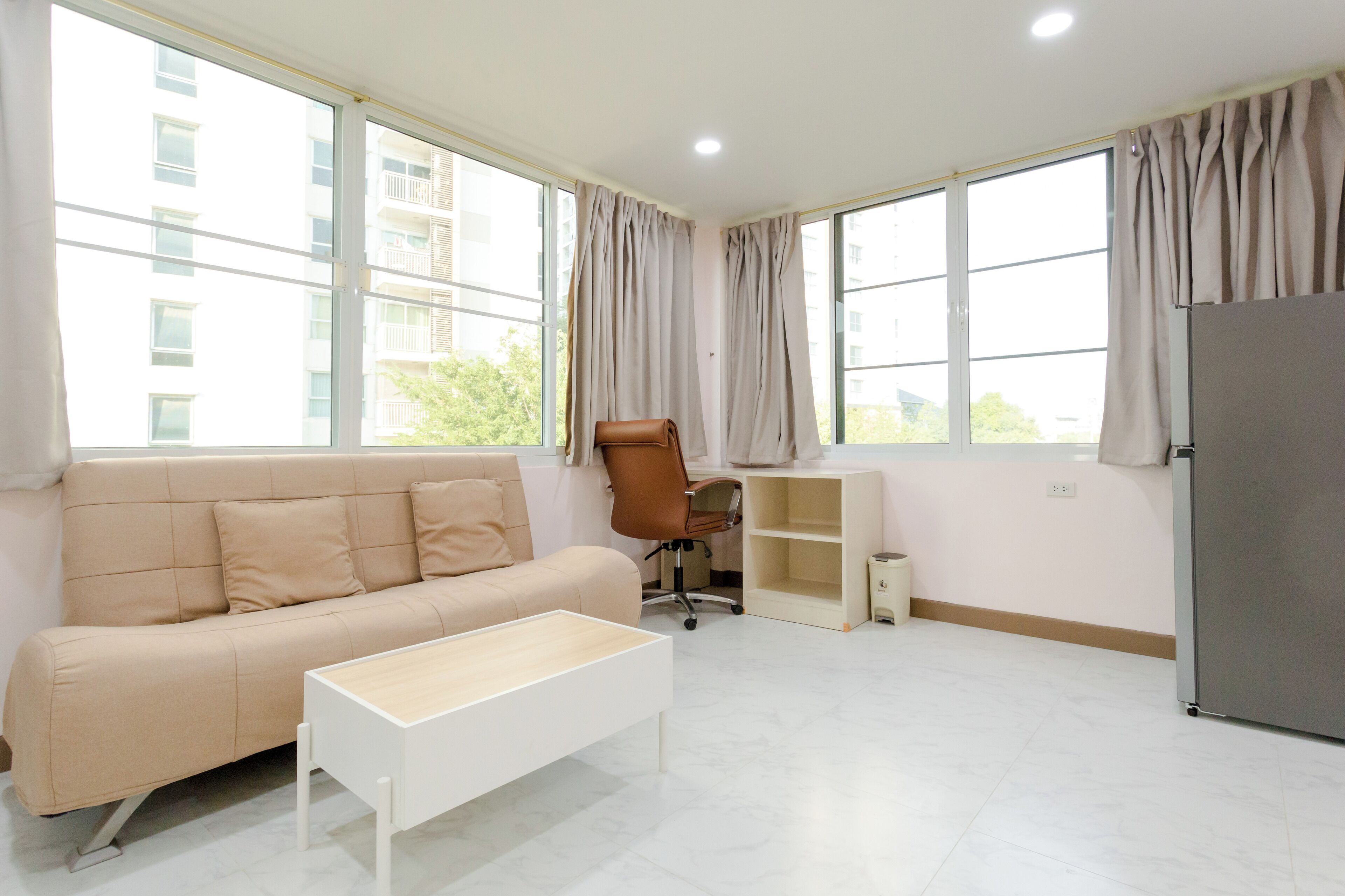 A modern living room with a beige sofa, a white coffee table, a brown office chair, a small desk, and a refrigerator.