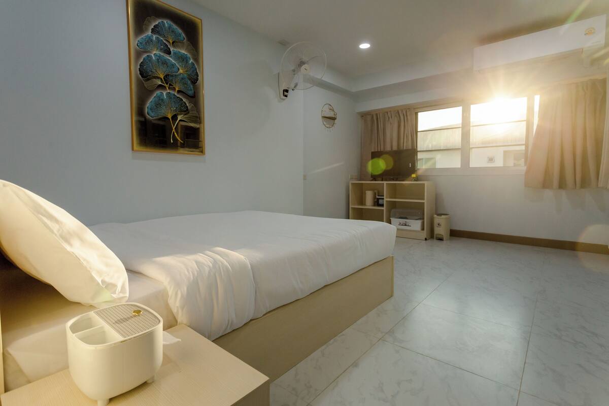 A bedroom with a bed, bedside table, lamp, and a wall-mounted fan.