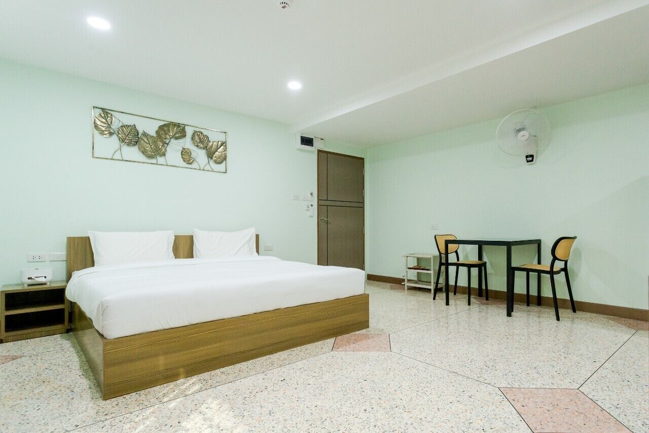 Une chambre d’hôtel avec un lit, un bureau avec deux chaises, un ventilateur fixé au mur et une porte.