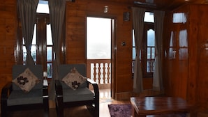 Interno di una baita in legno con due divani e un tavolo, ampie finestre con tende e un balcone con ringhiera.