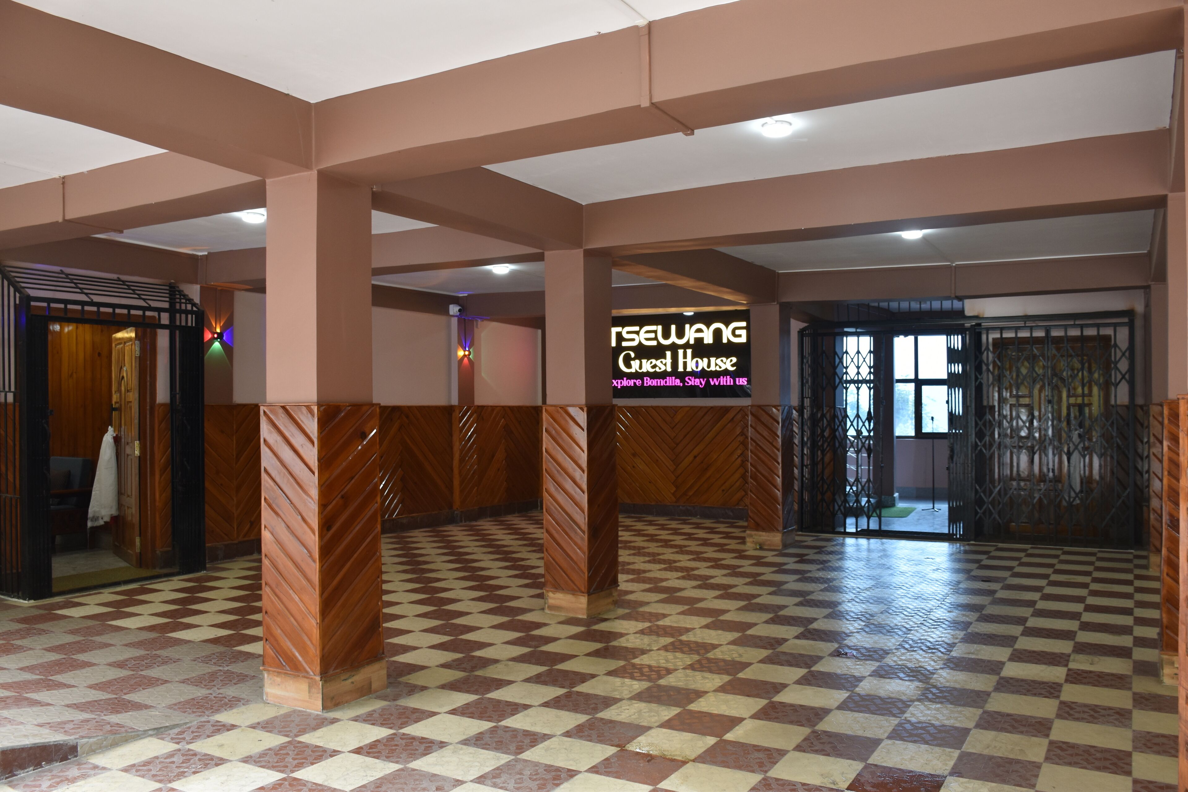 A hotel lobby with a checkered floor, wooden pillars, and a sign reading "RSEWANG Guest House."