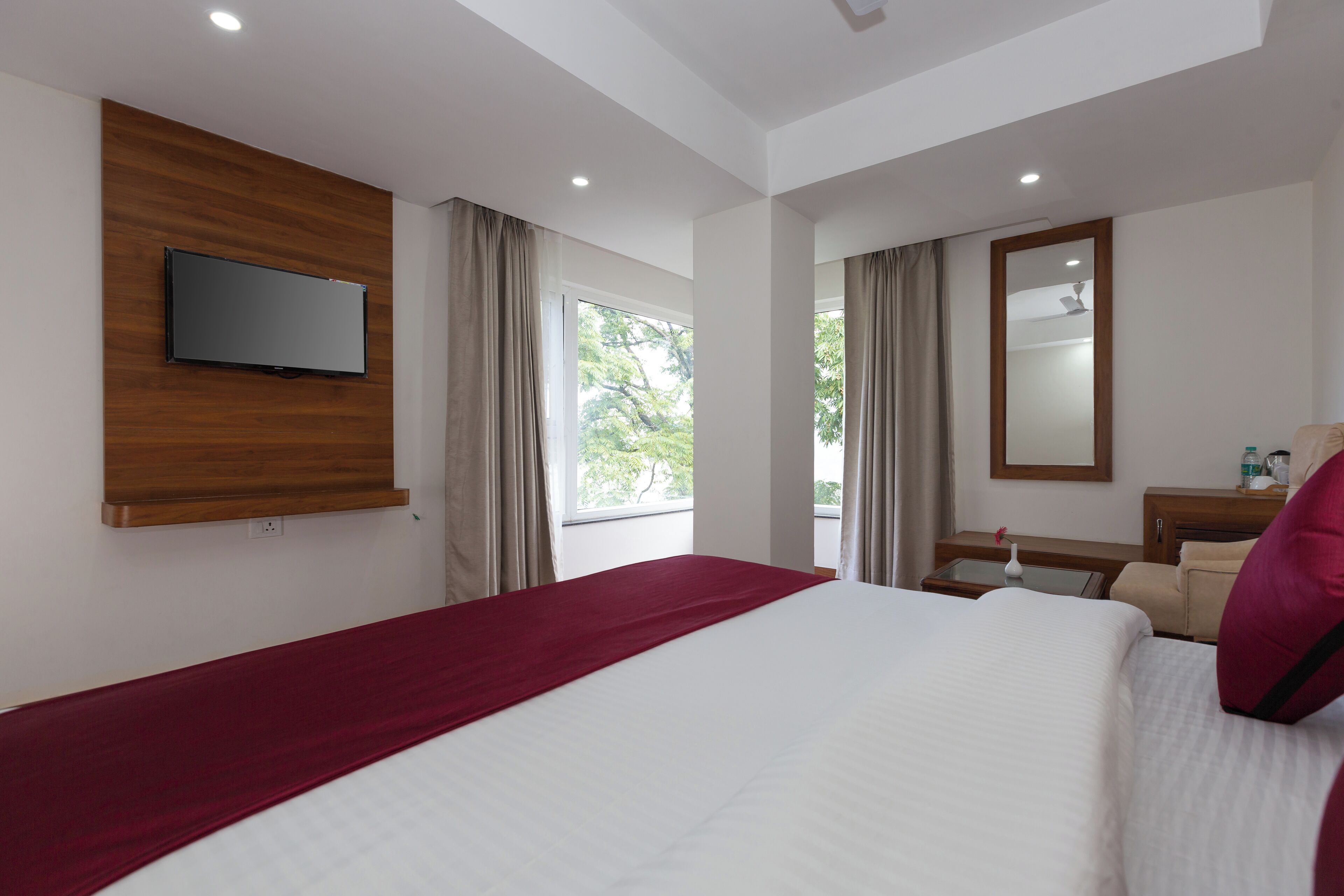 Ein Hotelzimmer mit einem Bett, einem an der Wand befestigten Fernseher, einem Fenster mit Vorhängen, einem Spiegel und einem kleinen Tisch mit einer Lampe.