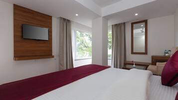 Ein Hotelzimmer mit einem Bett, einem an der Wand befestigten Fernseher, einem Fenster mit Vorhängen, einem Spiegel und einem kleinen Tisch mit einer Lampe.