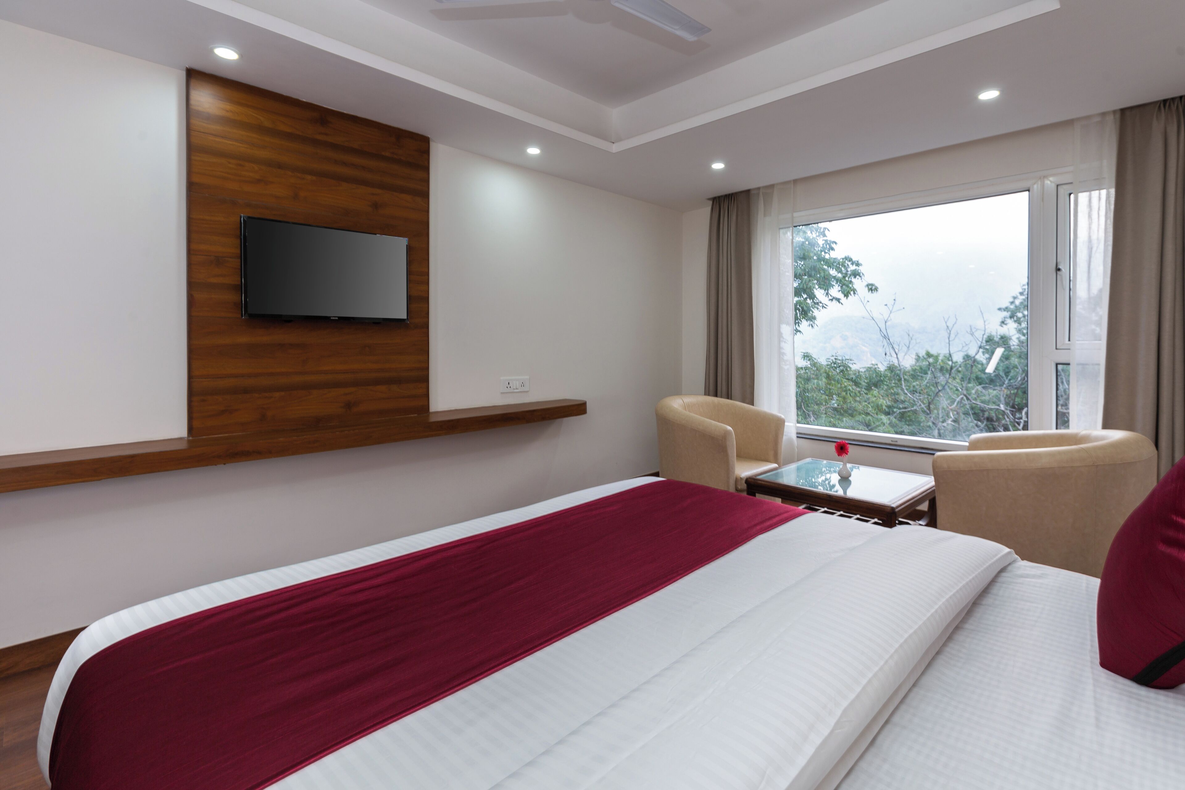 Quarto de hotel com cama, televisão presa na parede, janela com cortinas e uma cadeira.