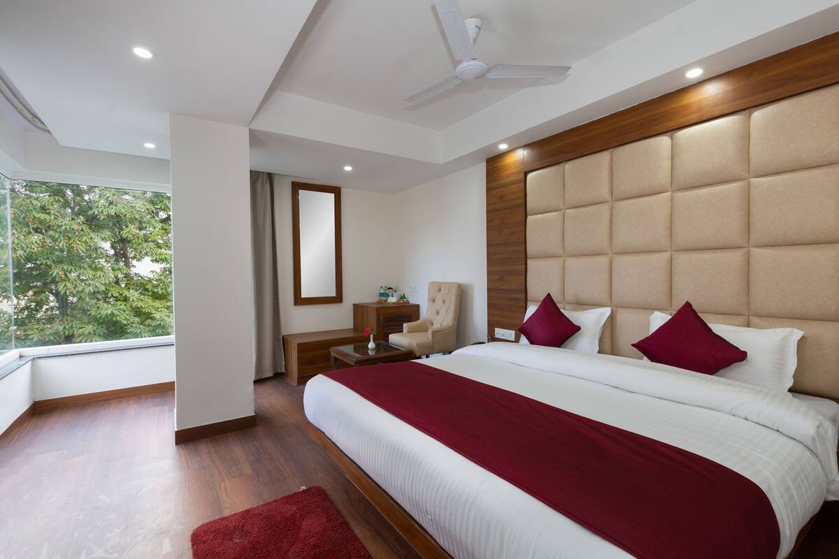A modern hotel room with a large bed, a ceiling fan, a wooden headboard, and a view of trees through the window.