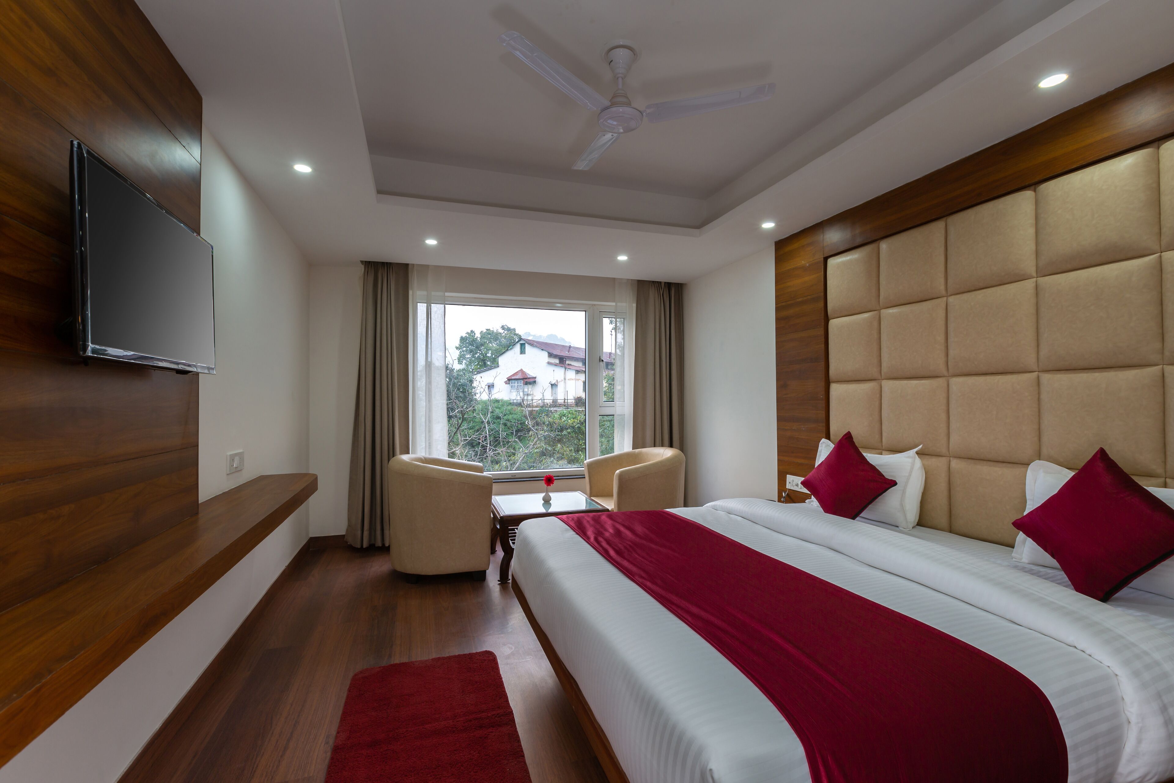A hotel room with a bed, a TV, a ceiling fan, a window with curtains, and a small table with chairs.