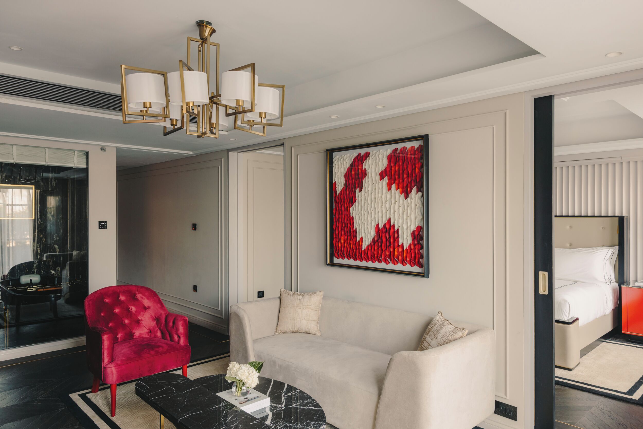Une chambre d’hôtel moderne avec un fauteuil rouge, un canapé beige, une table basse en marbre et une grande œuvre d’art abstraite accrochée au mur.