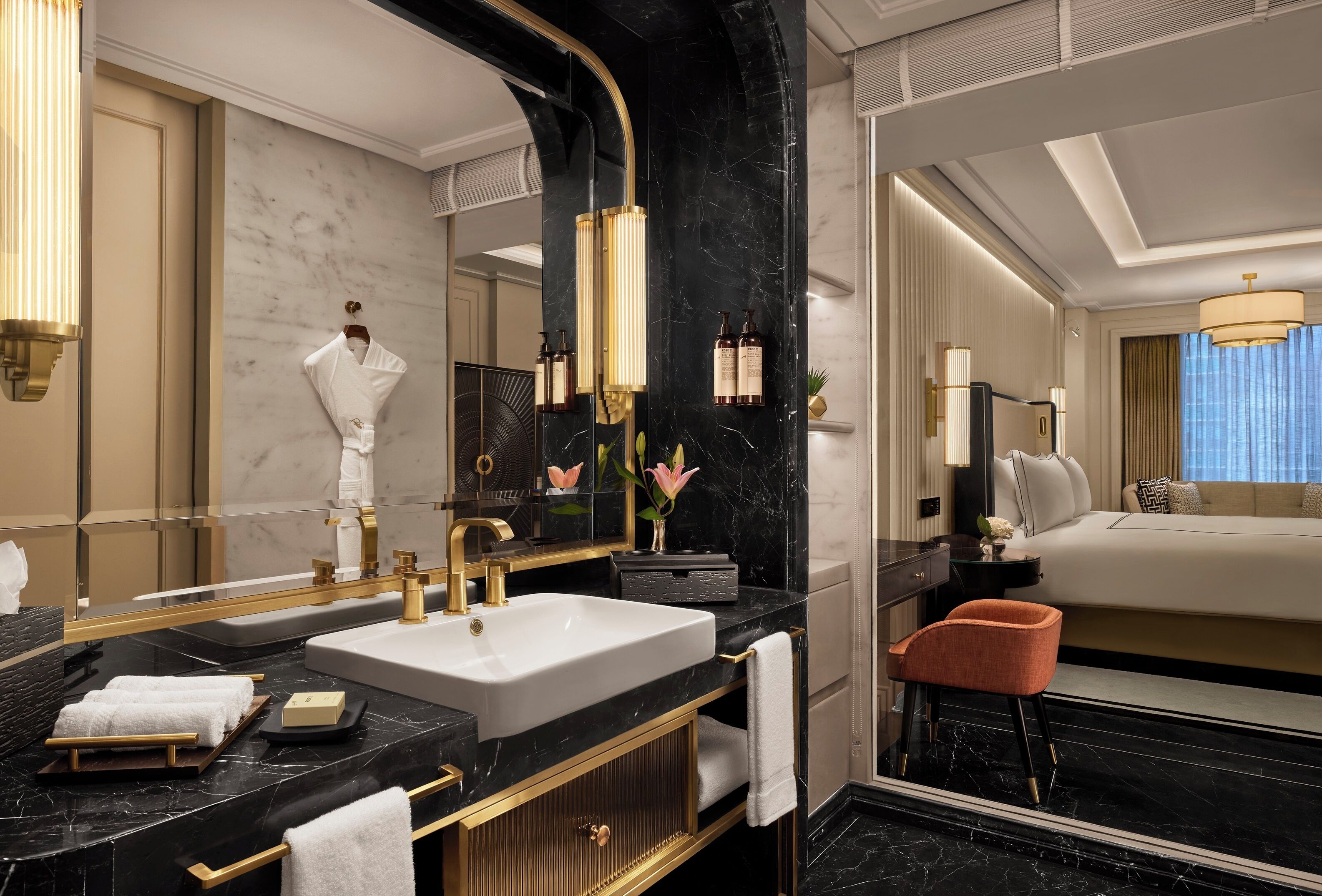 A luxurious bathroom with a large mirror, gold fixtures, a marble countertop, and a view of a bedroom with a bed and a nightstand.