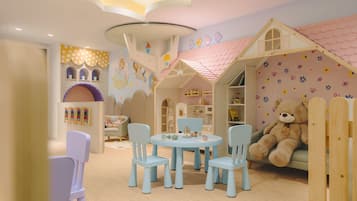 Um espaço infantil com uma casinha, um urso de pelúcia grande e uma mesa com cadeiras.