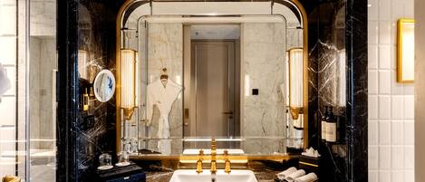 Un baño lujoso con un espejo grande, encimeras de mármol, un lavabo blanco y accesorios dorados.
