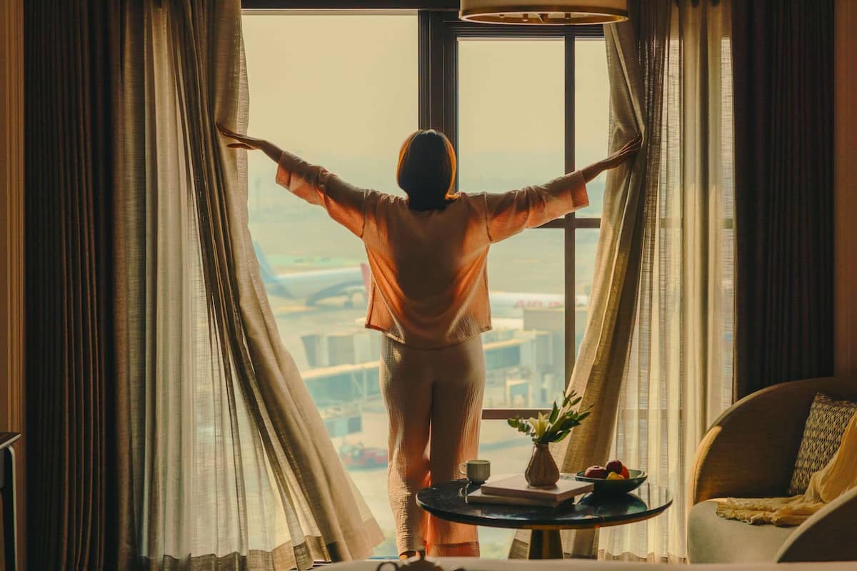 A person standing in a room with a view of a cityscape through a window, a table with a tray, and a chair.