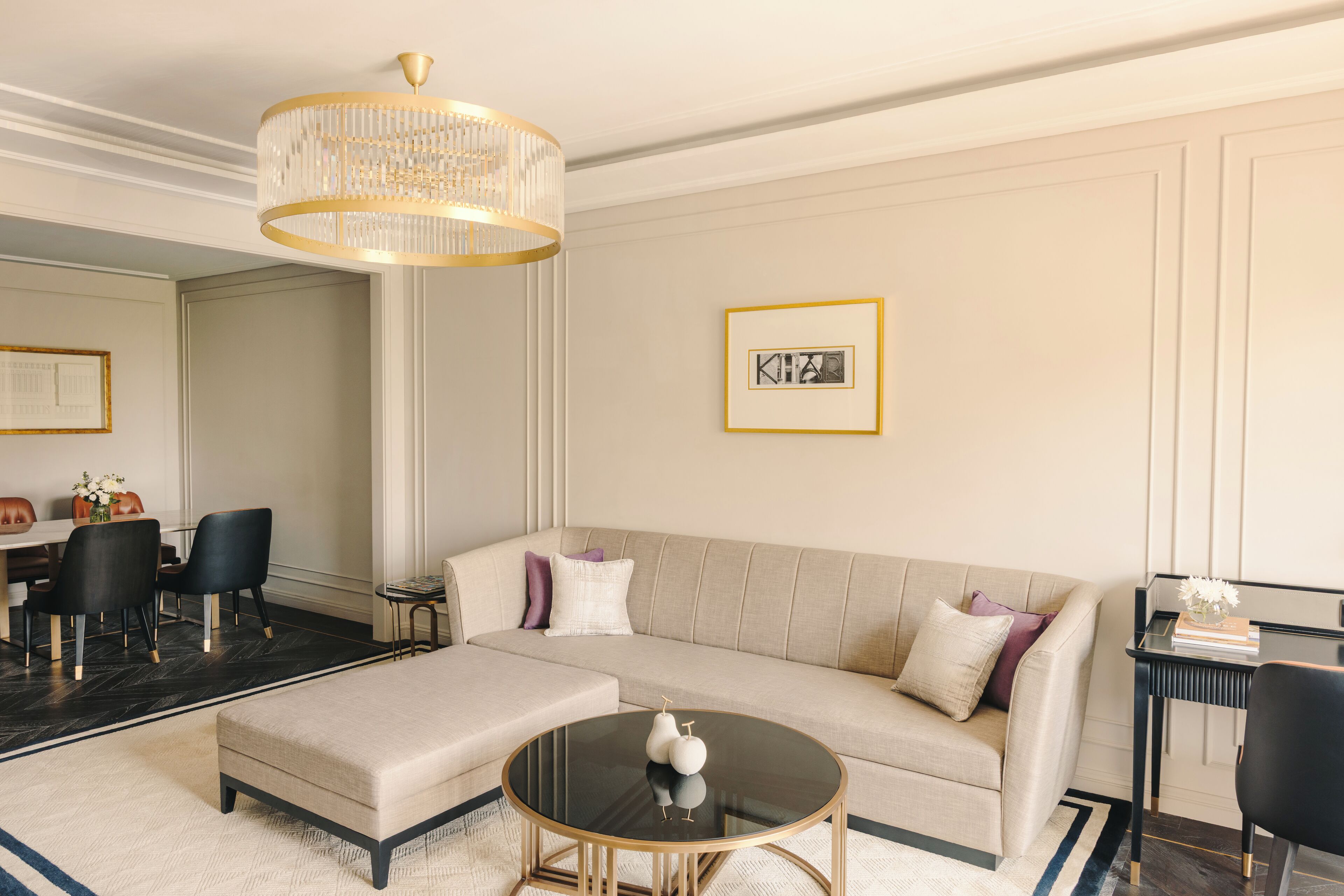 Una sala de estar moderna con un sofá seccional color beige, una mesa de centro redonda con objetos decorativos, una araña y un cuadro en la pared.