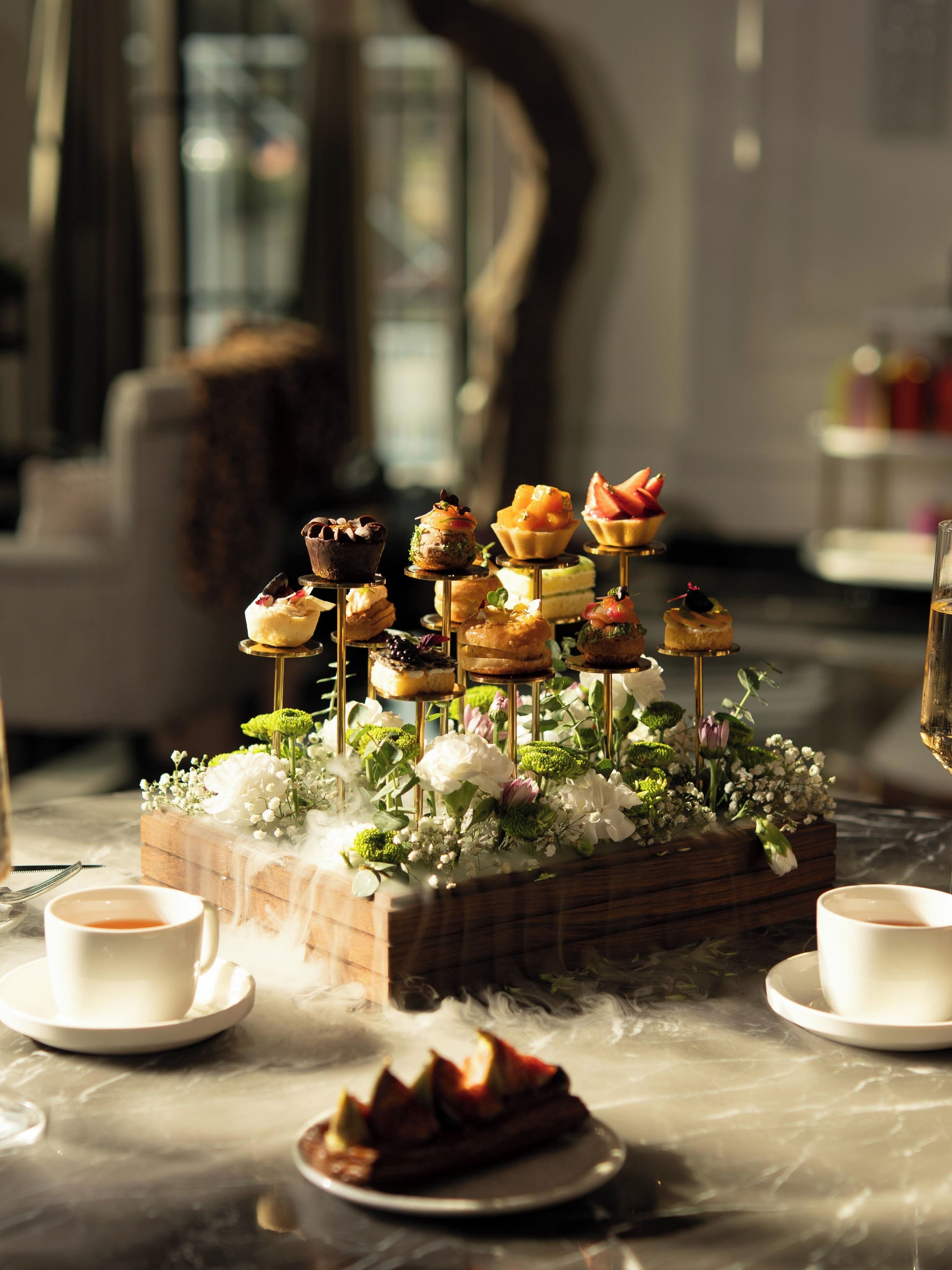 A dessert display with various pastries on stands, surrounded by white flowers and a wooden box.