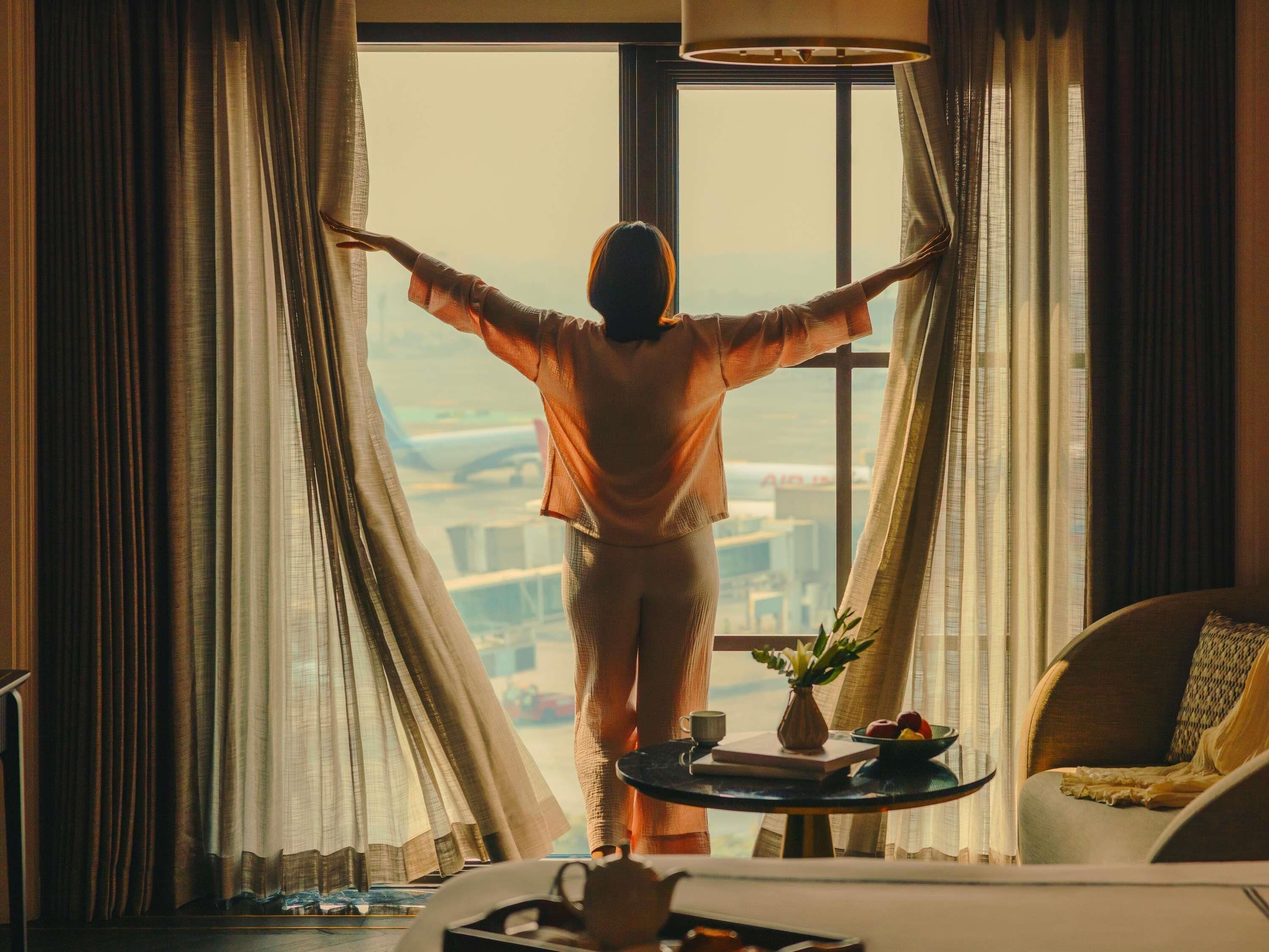 Una persona de pie en una habitación con vistas a un paisaje urbano a través de una ventana, una mesa con una bandeja y una silla.