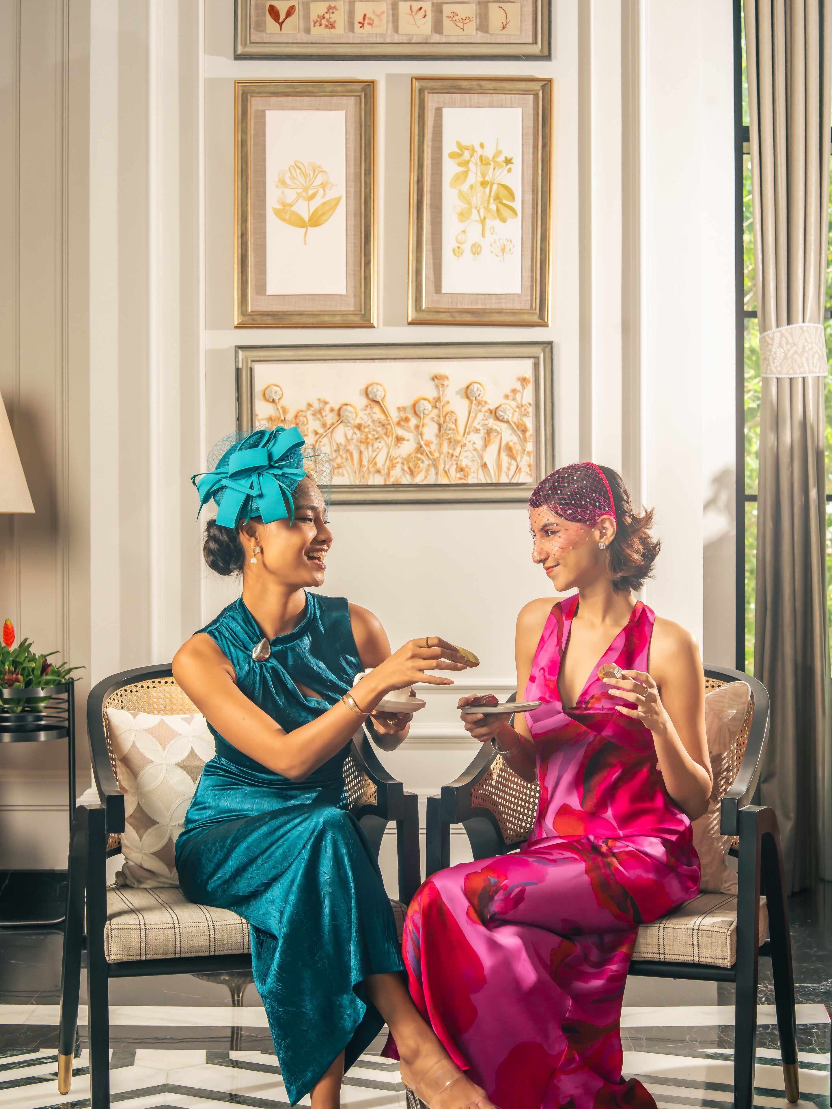 Two women seated on a sofa in a well-lit room with framed botanical artwork on the wall.