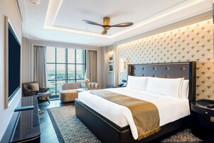 Habitación de hotel con una cama grande, una zona de estar con sofá y sillones, un escritorio con lámpara, y vistas de la ciudad a través de amplios ventanales.