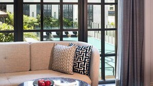 Habitación de hotel moderna con sofá, una mesa redonda con manzanas y vistas a un edificio y follaje a través de ventanas amplias.