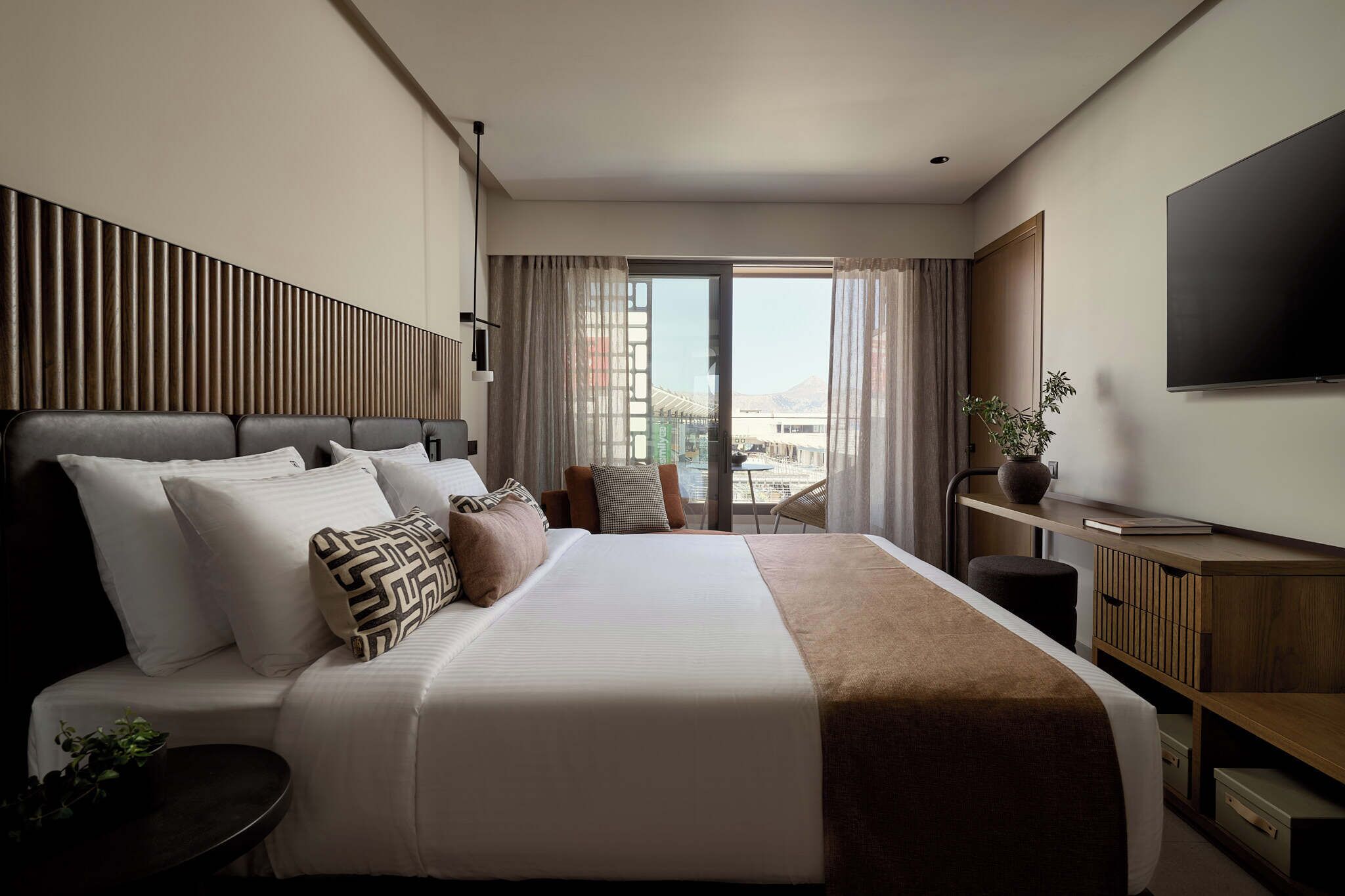 A modern hotel room with a large bed, a flat-screen TV, a desk with a chair, and a view of the city through large windows.